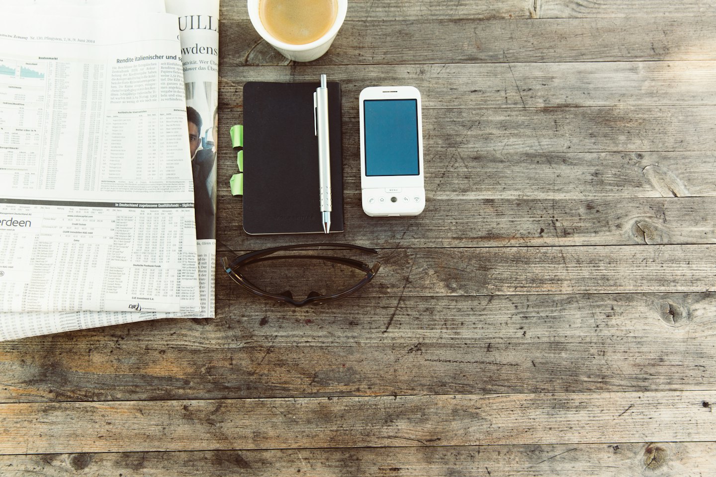 Oggetti da scrivania tra cui un giornale, una tazza di caffè, un taccuino e un telefono su un tavolo di legno.
